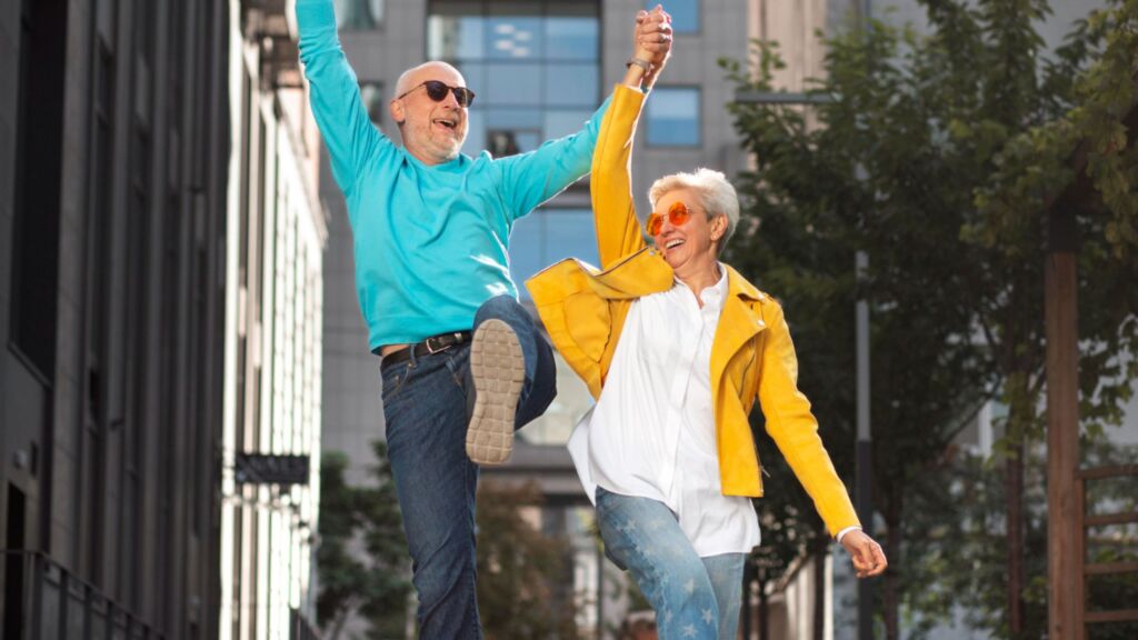 Older man in blue shirt and jeans holding hands with older woman in white shirt, yellow jacket, and jeans. The couple is jumping in the air and smiling with a city street in the background.
