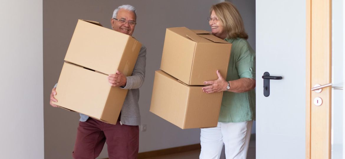 smiling-elderly-couple-moving-into-new-house-retired-husband-wife-holding-cardboard-boxes-downsizing