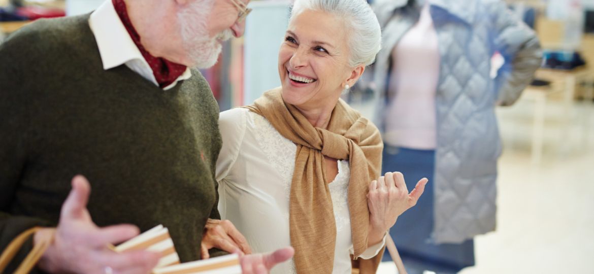 Smiling senior couple shopping together in a lively retail area, highlighting the benefits of a well-located senior living community with easy access to amenities.